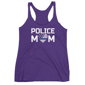 May include: A purple racerback tank top featuring the white distressed text POLICE MOM. The letter O is replaced by a heart shape containing a white and blue American flag pattern with stars and a horizontal blue stripe across the center of the heart.