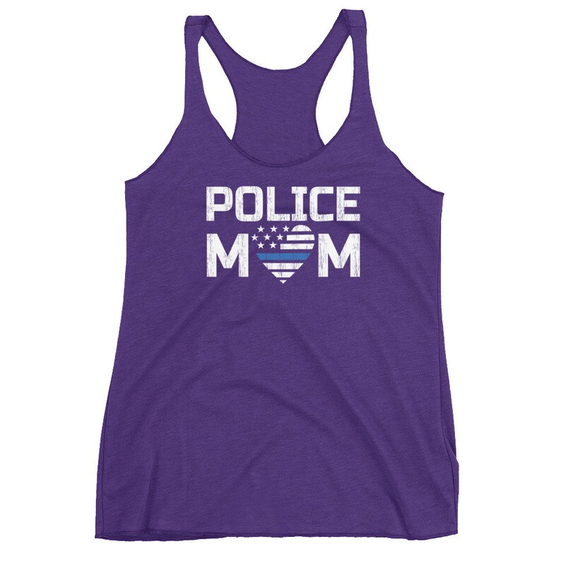 May include: A purple racerback tank top featuring the white distressed text POLICE MOM. The letter O is replaced by a heart shape containing a white and blue American flag pattern with stars and a horizontal blue stripe across the center of the heart.