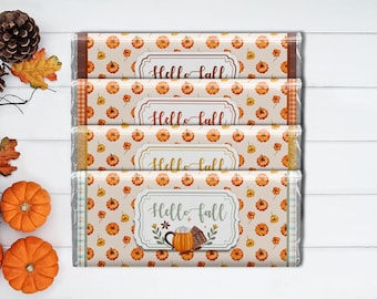 以下が含まれることがあります： 「Hello Fall」の文字が入った4本のチョコレートバー。それぞれオレンジ色のカボチャ模様の紙で包まれています。包装紙には、チェック柄やキラキラなど、さまざまなボーダーデザインがあります。白い木製の表面に配置されています。