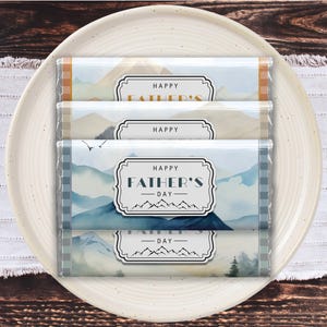 May include: Four chocolate bars with mountain landscape designs and the text "Happy Father's Day" on a white plate. The bars have blue, tan, and green hues, with a mountain range illustration. The plate sits on a white and beige patterned surface.