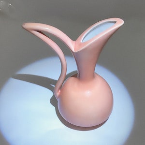 May include: A pink ceramic pitcher with a rounded body and a curved spout. The pitcher is sitting on a white surface.