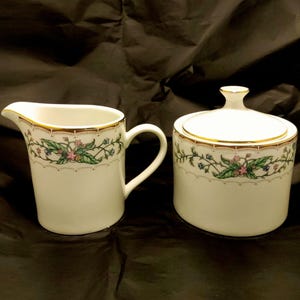 May include: A white porcelain creamer and sugar bowl set with a floral design and gold trim. The creamer has a handle and the sugar bowl has a lid.
