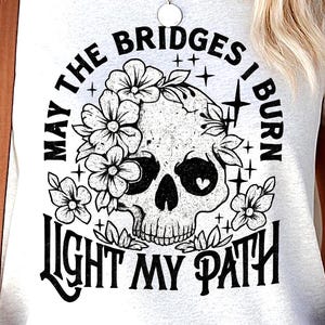 May include: White t-shirt with a skull design, floral accents, and the text "May the bridges I burn light my path." The graphic is in black and white. The shirt has short sleeves and a crew neck.
