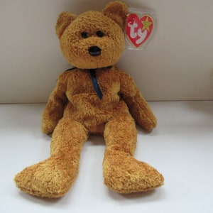 May include: A brown plush teddy bear with a blue ribbon around its neck. The bear has a Ty tag attached to its ear.