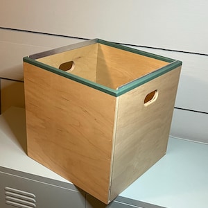 Puede incluir: Una caja de almacenamiento cuadrada de madera con un acabado de madera clara y un borde verde alrededor del borde superior. La caja tiene dos asas recortadas en el lateral.