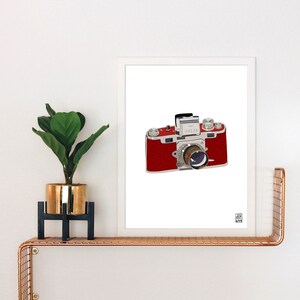 May include: A white framed print of a red and silver vintage camera with a flash attachment. The camera is on a white background.