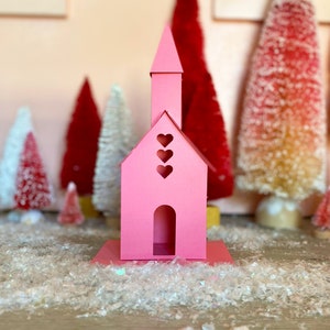 May include: A pink papercraft church with three heart cutouts on the front and a single heart cutout on the top. The church is sitting on a white surface with white glitter.