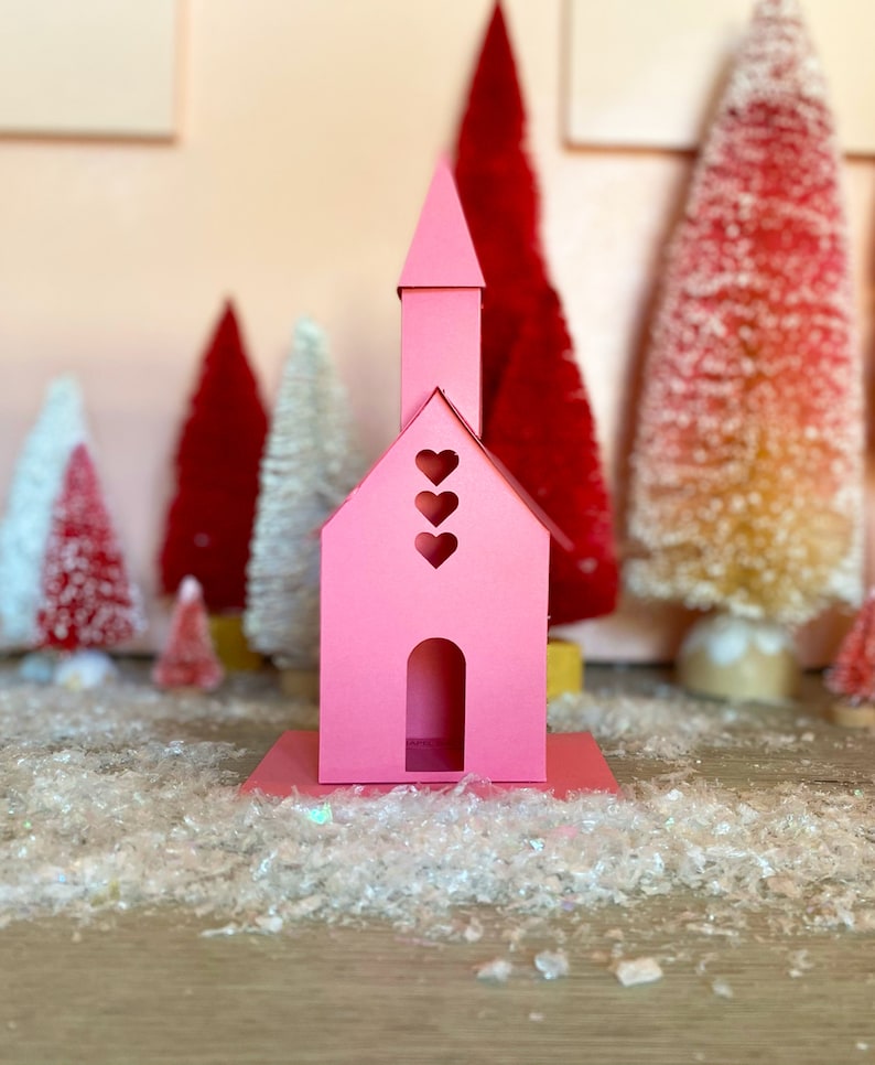 May include: A pink papercraft church with three heart cutouts on the front and a single heart cutout on the top. The church is sitting on a white surface with white glitter.