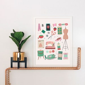 May include: A white framed print with a pink background featuring a collection of sewing and crafting supplies. The print includes a sewing machine, buttons, thread, scissors, a measuring tape, a dress form, and a vintage iron.