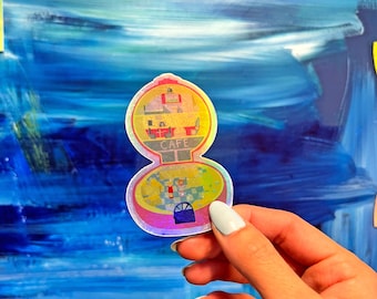 May include: A holographic sticker of a cafe building with a pink roof and a sign that says "Cafe". The sticker is on a blue background.