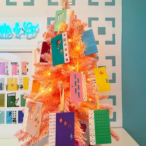 May include: A pink Christmas tree decorated with colorful door-shaped ornaments. The ornaments are various colors, including green, blue, yellow, and purple, with white decorative trim. The tree is lit with small, warm white lights, creating a festive display.