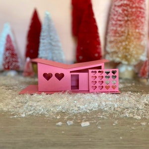 May include: A pink papercraft model of a mid-century modern house with heart-shaped windows and a fence. The house is sitting on a bed of white glitter.