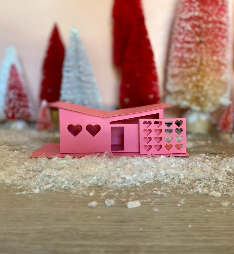 May include: A pink papercraft model of a mid-century modern house with heart-shaped windows and a fence. The house is sitting on a bed of white glitter.