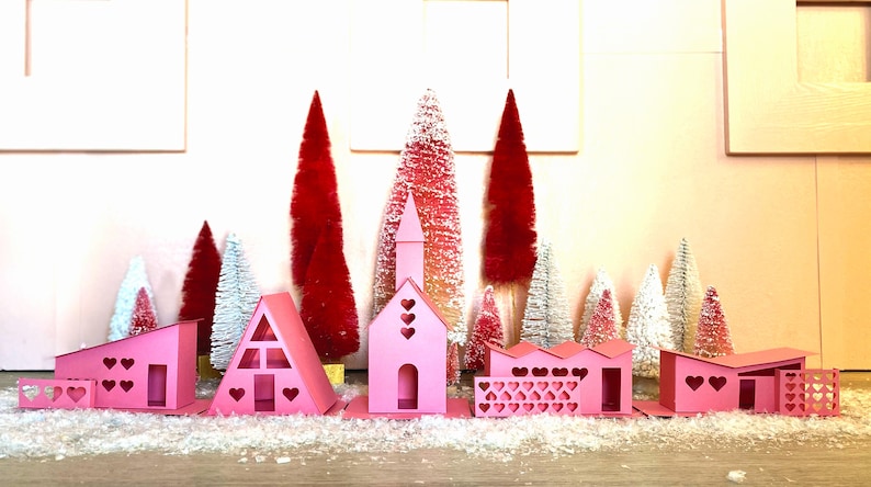 May include: A pink papercraft village with heart-shaped cutouts, surrounded by miniature pink and white flocked trees. The village includes a church, a house with a triangular roof, and a house with a flat roof.