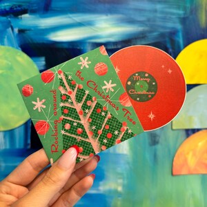 May include: A green and red Christmas card with a stylized Christmas tree and the text "Rocking around the Christmas Tree". The card is being held in a hand.