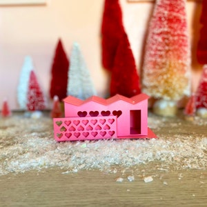 May include: A pink papercraft house with a heart-shaped fence and a small door. The house is sitting on a white, sparkly surface.