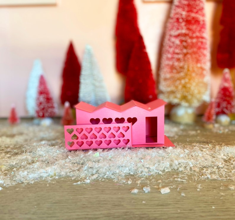 May include: A pink papercraft house with a heart-shaped fence and a small door. The house is sitting on a white, sparkly surface.