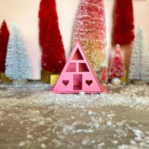 May include: A pink cardboard house shaped like a triangle with two heart cutouts and a window. The house is sitting on a white surface with white fluffy material scattered around it.