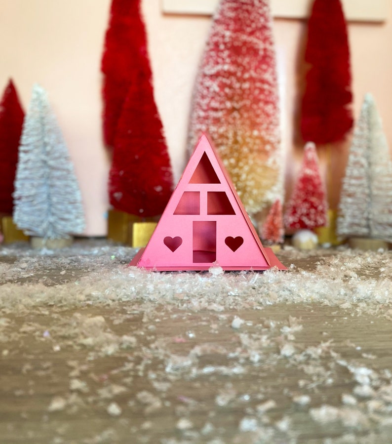 May include: A pink cardboard house shaped like a triangle with two heart cutouts and a window. The house is sitting on a white surface with white fluffy material scattered around it.