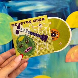 May include: A yellow vinyl record with the text "Happy Halloween" on it. The record is being held in a hand, and the cover art for the record is visible. The cover art features a green monster with a skull and crossbones and a guitar.