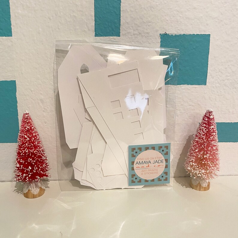 May include: A white paper house kit in a clear plastic bag. The kit includes multiple pieces to assemble a house. A small business card with the text "Find us on Etsy Amaya Jade and Co. Art Prints & Stickers" is included.