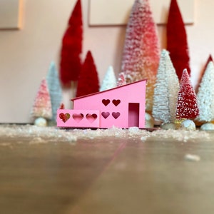 May include: A pink paper house with heart-shaped windows and a porch. The house is surrounded by miniature white and red Christmas trees.