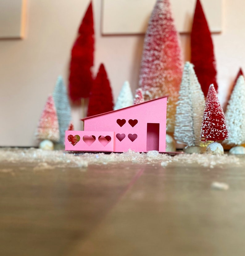 May include: A pink paper house with heart-shaped windows and a porch. The house is surrounded by miniature white and red Christmas trees.