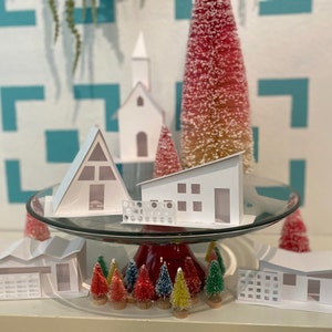 May include: A white papercraft village scene with a church, a-frame house, and a modern house. The houses are on a glass cake stand with a red base. There are small, flocked Christmas trees in front of the houses.