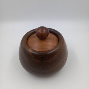 Puede incluir: Una caja de madera con tapa. La caja es de madera oscura y tiene forma redonda. La tapa es de madera más clara y tiene un pequeño pomo en la parte superior.