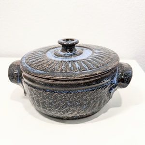 May include: A small, blue and brown ceramic pot with a lid. The pot has a textured surface and two handles. The lid has a raised design in the center.