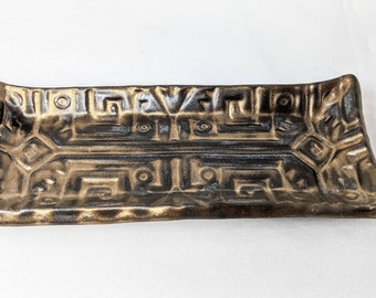 May include: A rectangular ceramic dish with a geometric design in gold and black. The dish has a raised rim and a slightly curved shape.