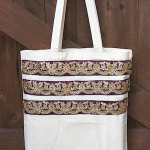 May include: A white canvas tote bag with three rows of gold and purple trim. The trim is a decorative ribbon with gold sequins and a purple base.