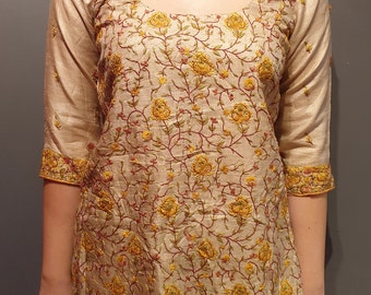 Puede incluir: Una túnica de seda beige con un patrón de bordado floral en tonos de amarillo, rojo y verde. La túnica tiene un escote redondo y mangas tres cuartos.