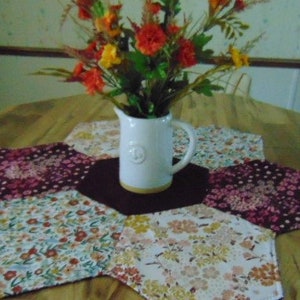 May include: A white ceramic pitcher with a handle holds a bouquet of orange and yellow flowers. The pitcher sits on a dark brown hexagonal mat, surrounded by six floral patterned, hexagonal placemats on a wooden table.