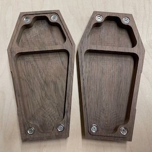 May include: Two wooden coffin-shaped boxes with a dark brown finish. Each box has a rectangular cutout in the center and is secured with four silver screws.
