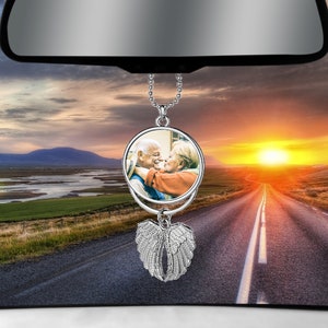 May include: A silver car charm featuring a photo of an older couple kissing. The charm hangs from a rearview mirror and has a pair of silver angel wings.