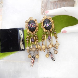 May include: A pair of Diana Marco dangle earrings featuring oval black cabochons with multicolored glitter accents. The gold-tone metal settings are adorned with hanging chains, textured gold beads, translucent purple beads, and speckled black and orange charms.