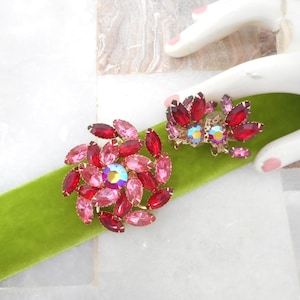 May include: Two vintage rhinestone brooches. One is a large, round brooch with pink and red stones surrounding an iridescent center stone. The other is a smaller, matching brooch with red and pink stones. Both are set on a green velvet ribbon.