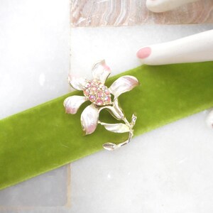 May include: A floral brooch with a silver-tone finish, featuring a cluster of pink rhinestones at the center and delicate white and pink petals. The brooch is set on a vibrant green velvet ribbon.