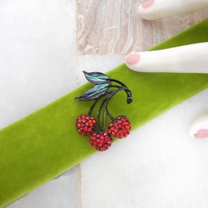 May include: A cherry brooch featuring three round red crystal-encrusted fruits attached to a black metal stem with two dark green enameled leaves. The brooch is positioned on a bright green velvet ribbon, showcasing a detailed fruit design.