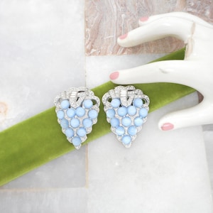May include: A pair of silver-toned clip-on earrings shaped like grape clusters. Each earring features round, light blue cabochon stones arranged in a triangular pattern, accented with small, clear rhinestones along the top and edges.