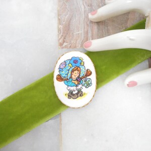 May include: An oval-shaped brooch featuring a cartoon illustration of a character with brown hair wearing a blue bonnet and blue clothing, holding a small green butterfly. The brooch is centered on a strip of bright green velvet ribbon.