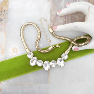 May include: A gold-toned necklace with clear, oval-shaped crystals. The necklace has a flexible, ribbed design and is displayed on a bright green velvet ribbon. The background is a white and gray marble surface.