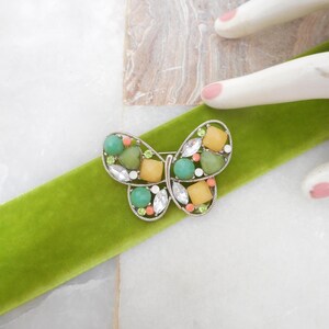 May include: A silver-toned butterfly brooch featuring a variety of decorative elements including round green beads, square yellow stones, clear marquise-cut crystals, and small coral-colored accents set within a delicate metal wire frame.