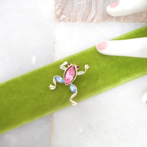 May include: A gold-toned frog brooch with a large pink center stone and blue and red accent stones. The brooch is set on a bright green velvet ribbon, with white ceramic hands in the background.