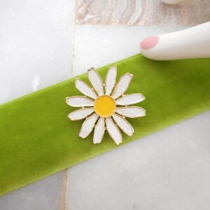 May include: A daisy brooch with white petals and a yellow center, set against a vibrant green velvet ribbon. The brooch has a gold-colored metal outline. The background is a white marble surface.