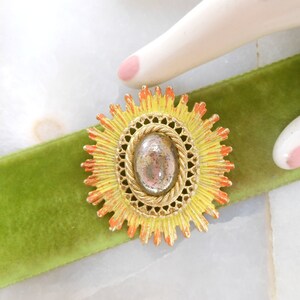 May include: A gold-toned brooch with a yellow and orange sunburst design. The center of the brooch features a round, gray, iridescent stone.
