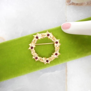 May include: A gold-toned wreath brooch with red accents, resting on a vibrant green velvet ribbon. The brooch features a circular design with holly leaf details and small red gems. The background is a white marble surface.