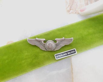 May include: A silver-colored military pilot's wing pin with a central emblem and outstretched wings. The pin is displayed on a bright green velvet ribbon, with a small "STERLING SILVER" tag nearby.