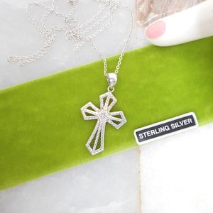 May include: A silver necklace with a cross pendant. The cross is detailed with a diamond-like center and a delicate openwork design. The necklace rests on a green velvet surface, with a "STERLING SILVER" tag.
